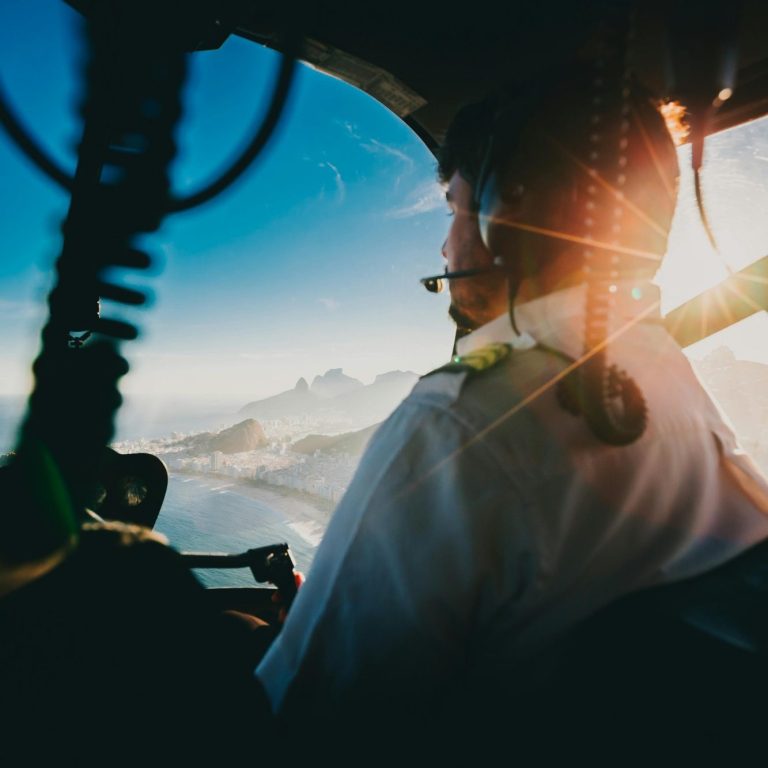 Un pilote d'hélicoptère en tenue professionnelle pris par un passager arrière. Un coucher de soleil est visible ainsi qu'un bord de mer.